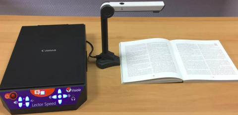 A thin magazine-type book on a table and under a pedestal-style scanner. The scanner is connected to a device that resembles a standard flatbed scanner with large, touch button controls on the inclined, front side. 