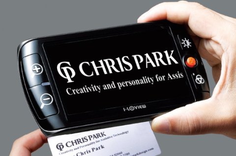 A black cell phone-like device with a black screen showing text enlarged from a business card below it.